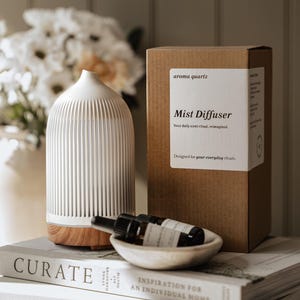 May include: A white, vertically ribbed aroma quartz mist diffuser with a wood-grain base, positioned next to a brown cardboard box labelled Mist Diffuser, and a small white bowl containing two dark glass dropper bottles, all resting on stacked books.