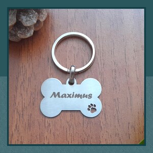 May include: A silver-colored dog tag in the shape of a bone, attached to a keyring. The tag has the name "Maximus" engraved on it, along with a paw print. The tag is displayed on a wooden surface.