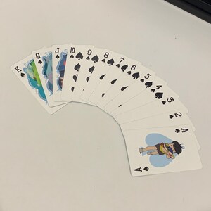 May include: A fan of playing cards featuring the suits of spades and hearts. The cards display the numbers 2 through 10, Jack, Queen, and King of Spades. The Ace of Hearts features an illustration of two characters. The cards are white with black symbols.