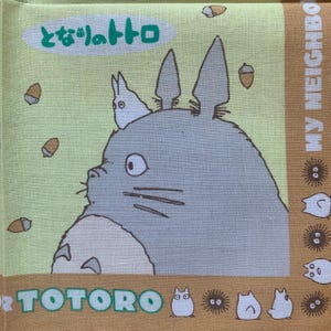 K&ouml;nnte beinhalten: Quadratischer, hellgr&uuml;ner und beigefarbener Stoff mit der Figur Totoro aus dem Zeichentrickfilm Mein Nachbar Totoro. Das Bild enth&auml;lt den japanischen Text "となりのトトロ" und das Wort "TOTORO". Eicheln und andere Charaktere sind ebenfalls zu sehen.