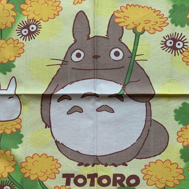 May include: A light yellow fabric featuring the character Totoro from the animated film. Totoro is a large, gray forest spirit holding a yellow flower. The word "TOTORO" is printed in brown at the bottom. The background has yellow flowers and other characters.