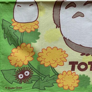 May include: A green and yellow patterned fabric featuring a cartoon character and dandelions. The design includes a large, round character with a white face and brown outline, along with yellow dandelions and green leaves. The text "TOT" is visible.