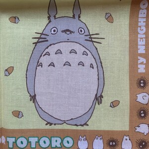 K&ouml;nnte beinhalten: Ein hellgr&uuml;ner und brauner Stoff mit einer gro&szlig;en, grauen Totoro-Figur mit wei&szlig;em Bauch und Eicheldetails. Der Text "TOTORO" befindet sich unten, mit "MY NEIGHBOR" auf der rechten Seite.
