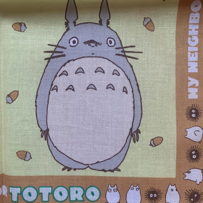 K&ouml;nnte beinhalten: Ein hellgr&uuml;ner und brauner Stoff mit einer gro&szlig;en, grauen Totoro-Figur mit wei&szlig;em Bauch und Eicheldetails. Der Text "TOTORO" befindet sich unten, mit "MY NEIGHBOR" auf der rechten Seite.