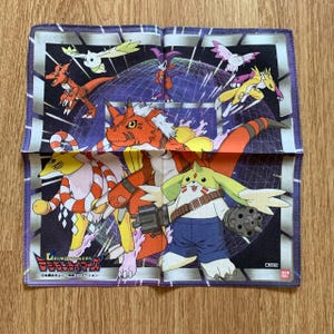 Peut inclure: Foulard Digimon carré et plié. Le motif présente divers personnages Digimon aux couleurs vives sur un fond violet et blanc. Les personnages Digimon sont dans diverses poses, certains tenant des armes.