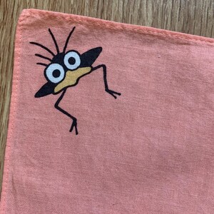May include: A coral-colored square handkerchief featuring a whimsical cartoon creature. The creature has a black body, large white eyes, and a yellow beak. Black lines form the legs and antennae.