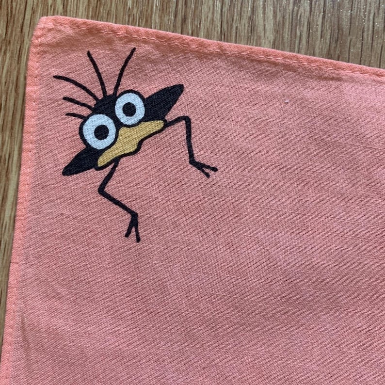 May include: A coral-colored square handkerchief featuring a whimsical cartoon creature. The creature has a black body, large white eyes, and a yellow beak. Black lines form the legs and antennae.