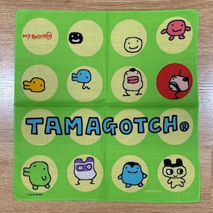 Op de afbeelding: Groene vierkante zakdoek met de tekst "TAMAGOTCH®" in blauw en cartoonfiguren in gele cirkels. De zakdoek heeft ook de tekst "MY GOTCH@" in rood.