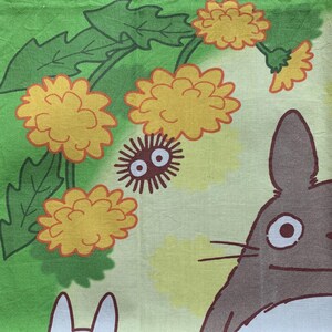 May include: A colorful fabric featuring a cartoon design. The design includes yellow flowers, green leaves, and a brown character with large eyes and whiskers. The background is a mix of green and yellow.