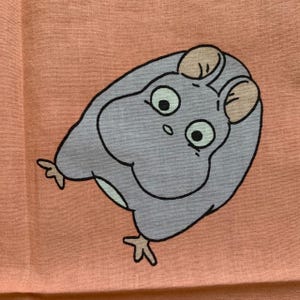 May include: A light purple cartoon character with large eyes and small ears is printed on a peach-colored fabric. The character has a round body and is outlined in black. The fabric has a slightly textured appearance.