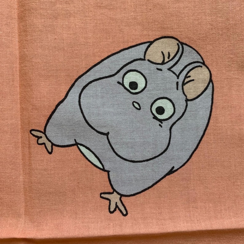 May include: A light purple cartoon character with large eyes and small ears is printed on a peach-colored fabric. The character has a round body and is outlined in black. The fabric has a slightly textured appearance.