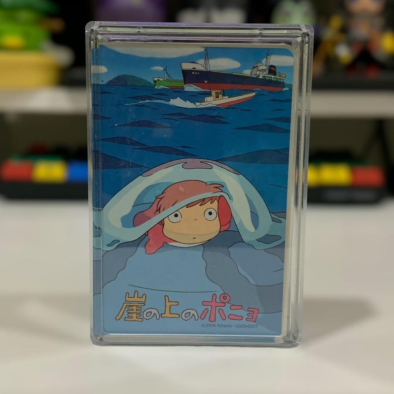 May include: A clear plastic case featuring a colorful illustration from the animated film "Ponyo." The artwork depicts a character with pink hair and a blue sea background with boats and ships. Japanese text is visible at the bottom.