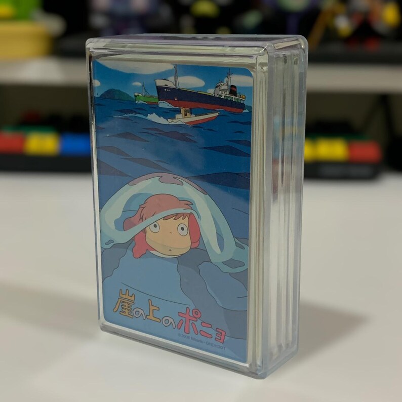 May include: A clear plastic case containing a deck of cards featuring an illustration from the animated film "Ponyo". The artwork depicts a character with pink hair and a blue sea background, with a ship and small boat in the distance. The Japanese title is visible.