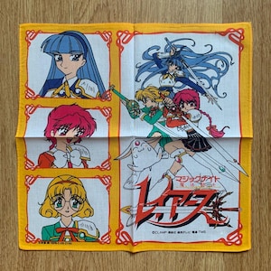 Puede incluir: Un pañuelo amarillo y blanco con ilustraciones de estilo anime. El diseño incluye varios personajes con espadas y un logotipo en japonés. El borde es amarillo con patrones decorativos.