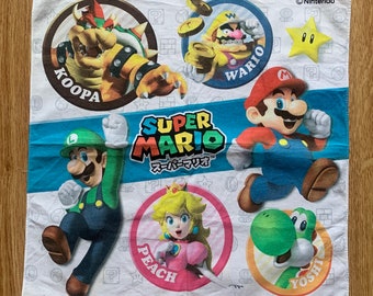 May include: A white cotton hand towel with a colorful Super Mario Bros. design. The towel features characters like Mario, Luigi, Peach, Yoshi, Bowser, and Wario. The text "SUPER MARIO" and "スーパーマリオ" is printed on the towel.