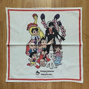 May include: White square cloth with a red border. It features a pixelated illustration of cartoon characters. The cloth has the text "TEZUKA OSAMU x TOKYO PIXEL" and copyright information.