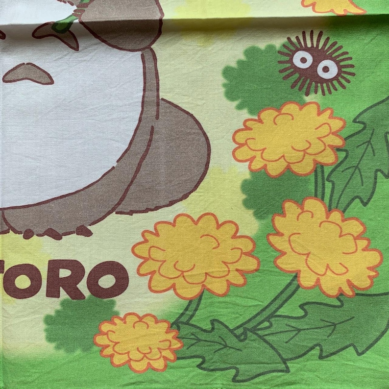 May include: A light green and yellow textile featuring a cartoon character with the word "TORO". The design includes yellow dandelions with green leaves and a small, round, black creature with white eyes.