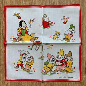 Op de afbeelding: Witte vierkante zakdoek met een rode rand met een Sneeuwwitje en de Zeven Dwergen ontwerp. De illustratie bevat Sneeuwwitje, herten, vogels en de dwergen in kleurrijke outfits. Het ontwerp is tegen een groen-witte achtergrond.