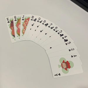 May include: A fan of playing cards featuring a unique design. The cards are white with black suit symbols and numbers. The face cards have colorful illustrations. The Ace of Clubs features a cartoon character. The cards are arranged in a semi-circle.