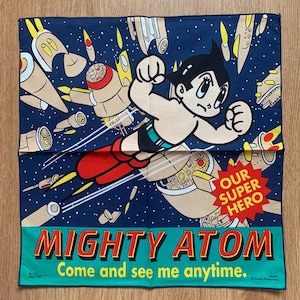 May include: A square fabric handkerchief featuring a dark blue space background with Astro Boy flying among rockets. A red starburst contains the text OUR SUPER HERO. The bottom teal border displays the text MIGHTY ATOM Come and see me anytime.