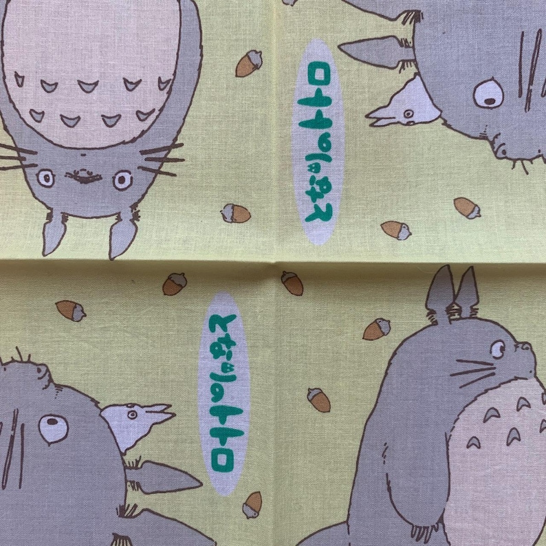 K&ouml;nnte beinhalten: Ein hellgelber Stoff mit Illustrationen der Figur Totoro aus dem Zeichentrickfilm Mein Nachbar Totoro. Das Design umfasst mehrere Totoro-Figuren, Eicheln und japanischen Text in einem gr&uuml;nen Oval.