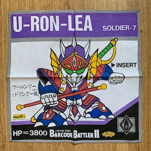 Puede incluir: Un paño cuadrado blanco y gris con una colorida ilustración de un guerrero robot con una espada y un bastón. El paño tiene el texto "U-RON-LEA" y "SOLDIER-7". El texto adicional incluye "BARCODE BATTLER II" y caracteres japoneses.