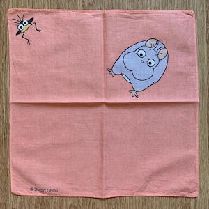 May include: A coral-colored handkerchief featuring two cartoon characters. One is a small, bug-like creature in black and the other is a light purple, hamster-like character. The words "Studio Ghibli" are printed in black at the bottom.