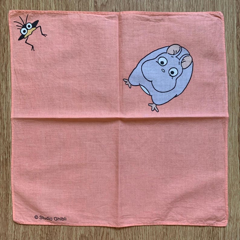May include: A coral-colored handkerchief featuring two cartoon characters. One is a small, bug-like creature in black and the other is a light purple, hamster-like character. The words "Studio Ghibli" are printed in black at the bottom.