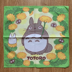 May include: A green square cloth featuring the character Totoro from the animated film. Totoro is depicted holding a dandelion, surrounded by yellow flowers and small, dark creatures. The word "TOTORO" is printed in brown.