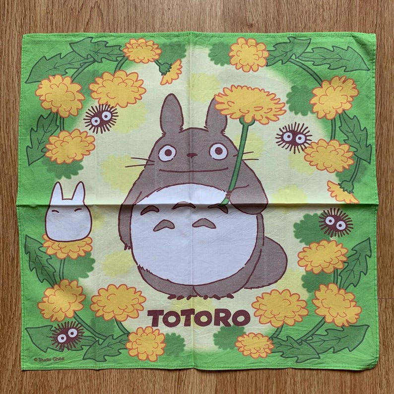 May include: A green square cloth featuring the character Totoro from the animated film. Totoro is depicted holding a dandelion, surrounded by yellow flowers and small, dark creatures. The word "TOTORO" is printed in brown.