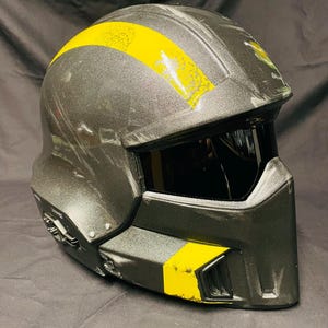 Może przedstawiać: Ciemnoszary kask motocyklowy z żółtym paskiem na górze i żółtym akcentem z boku. Kask ma przyciemniany wizjer i nowoczesny, kanciasty design. Kask jest przeznaczony do bezpieczeństwa i ochrony.