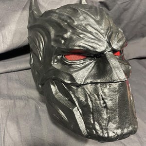 May include: A black, textured mask with pointed ears and red mesh eye coverings. The mask has a detailed, sculpted design and is likely for cosplay or costume use. The overall theme is superhero or comic book inspired.