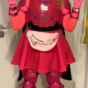 Op de afbeelding: Een persoon die een roze en rood kostuum draagt met een Hello Kitty-thema helm, borststuk en heuptas. Het kostuum bevat ook roze handschoenen en zwarte laarzen.