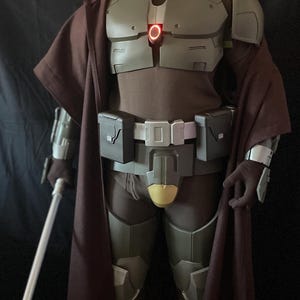 May include: A full-body shot of a costume featuring olive green and brown armor with a red illuminated chest piece. The costume includes a brown cape, gloves, and a belt with pouches. A white lightsaber is held in the left hand.