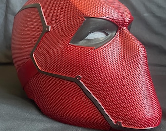 Può includere: Un casco integrale caratterizzato da una superficie rossa testurizzata con un motivo a punti ripetuti. Il casco presenta aperture oculari angolari scure con una rete e linee di pannelli rosso scuro lisce che delineano la calotta esterna.
