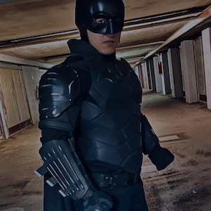 Può includere: Un costume da Batman nero, che include un cappuccio con orecchie a punta, una piastra pettorale e protezioni per le braccia. Il costume ha un aspetto strutturato e viene indossato con un mantello e pantaloni neri. Il tema generale è il cosplay.