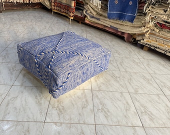 May include: A square blue pouf with white accents. The woven fabric has a textured appearance. White rope-like trim outlines the edges and forms a decorative cross pattern on the top. This floor cushion is a stylish home decor item.