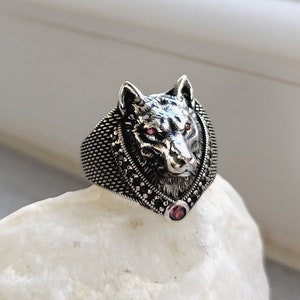 Könnte beinhalten: Ein silberner Ring mit einem Wolfskopf-Design und einem roten Edelstein. Der Ring liegt auf einem weißen Stein.