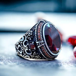 May include: A silver ring with an ornate design featuring a large, oval-shaped red gemstone surrounded by smaller red gemstones.