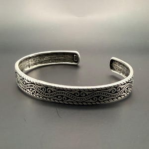 May include: A silver cuff bracelet with an intricate wave pattern. The open-ended bracelet has a textured finish and a detailed design that wraps around the entire band. The bracelet is displayed on a dark gray surface.
