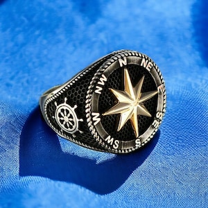 May include: A silver ring with a black compass face and a gold star. The compass face has the letters N, E, S, and W around the edge. The ring also has a small silver wheel on the side.