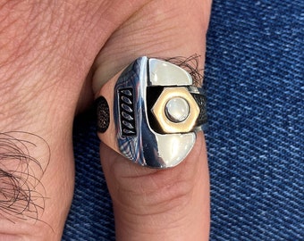 May include: A silver ring with a wrench design and a gold-colored nut. The ring is worn on a finger.