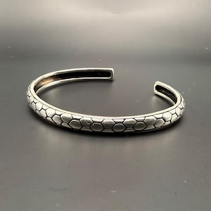 May include: A silver cuff bracelet with a textured, honeycomb pattern. The open-ended design features a series of connected hexagonal shapes, creating a geometric and modern aesthetic. The bracelet is displayed against a dark background.