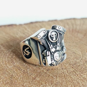 May include: A silver ring with a detailed motorcycle engine design. The engine has a skull in the center and the ring has a '13' engraved on the side.
