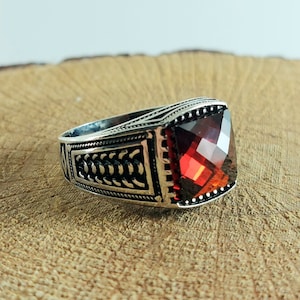 Puede incluir: Un anillo de plata con una piedra preciosa roja. El anillo tiene un diseño detallado con un patrón trenzado.