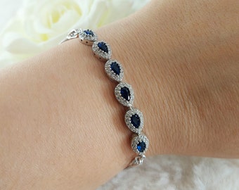 Op de afbeelding: Een zilveren armband met een rij van traanvormige blauwe stenen, omringd door kleinere heldere stenen.