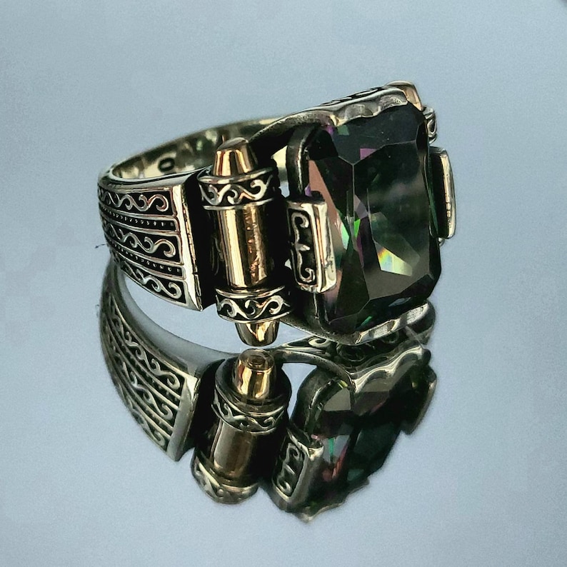 May include: A silver ring with a large, faceted, dark green gemstone. The ring has intricate, detailed designs on the band and a gold accent on the side.