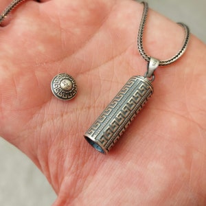 May include: A silver chain necklace with a silver and gold cylindrical pendant featuring a Greek key pattern. The pendant has a small silver disc with the number "925" on it.