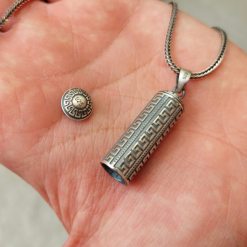 May include: A silver chain necklace with a silver and gold cylindrical pendant featuring a Greek key pattern. The pendant has a small silver disc with the number "925" on it.