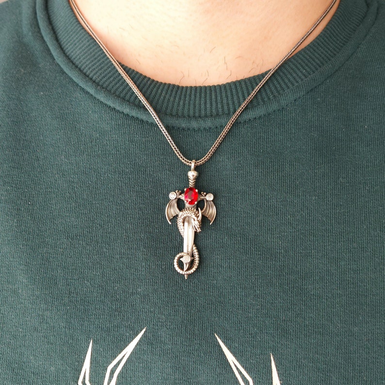 May include: A silver necklace with a pendant featuring a dragon wrapped around a sword with a red gemstone.